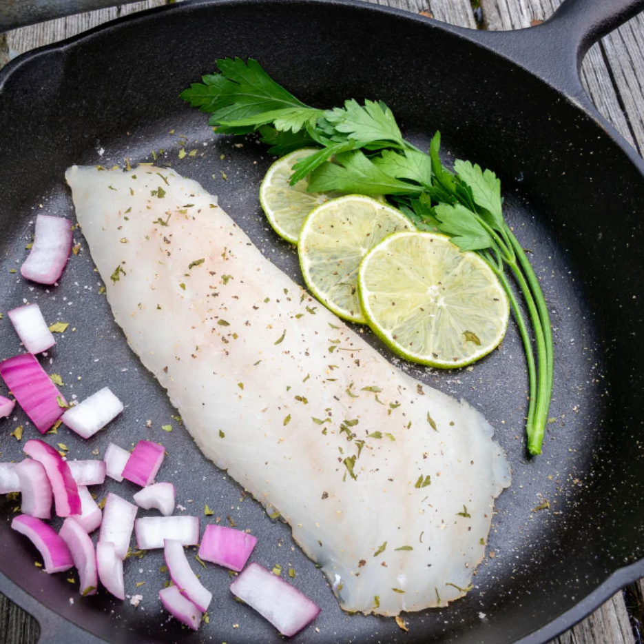 Raw fish fillet with lime slices and onions in a cast iron skillet on a wooden surface.